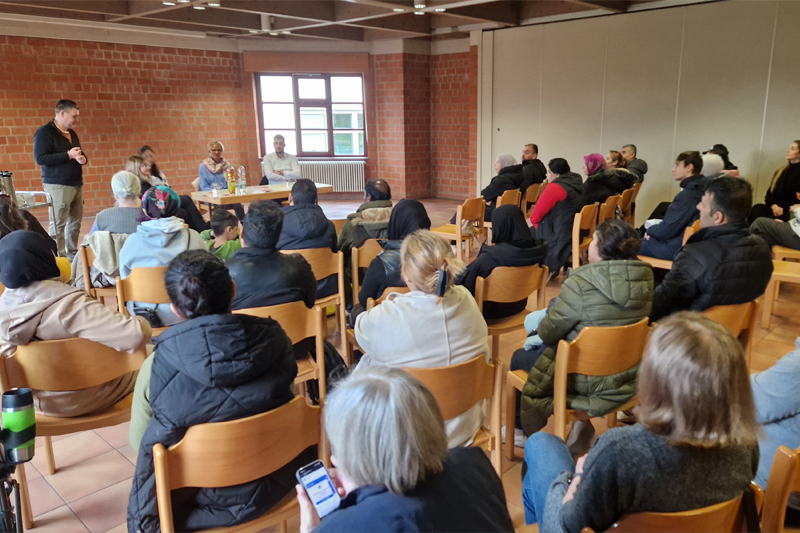 Rückansicht von Menschen, die auf Holzstühlen sitzen. Vor ihnen ein Mann, der spricht, sowie weitere Personen an einem Tisch.