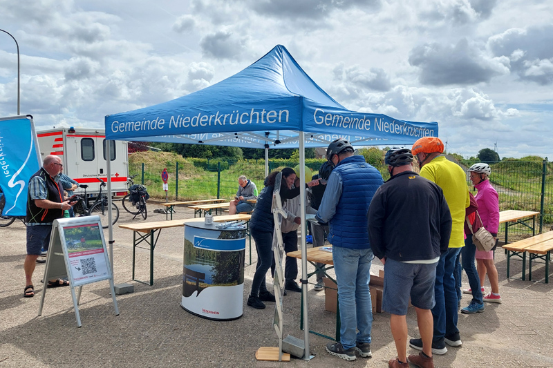 Der Infopunkt der Gemeinde Niederkrüchten beim Raderlebnistag. Ein blauer Pavillon mit Counter, vor dem Teilnehmende der Veranstaltung stehen und sich informieren.
