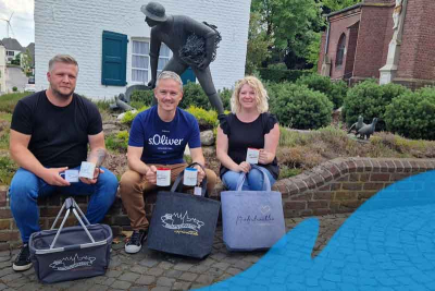 Oliver Greiss (Stoffjule), Frank Grusen (Gemeinde Niederkrüchten) und Julia Greiss (Stoffjule) mit Stofftaschen, einem Einkaufskorb und Tassen mit dem Schriftzug "Niederkrüchten"