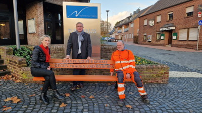 Christiane Jung, Gleichstellungsbeauftragte der Gemeinde Niederkrüchten, Bürgermeister Karl-Heinz Wassong und Bernd Pross vom Bauhof der Gemeinde auf einer orangenen Bank mit der Aufschrift "Kein Platz für Gewalt gegen Frauen und Mädchen"