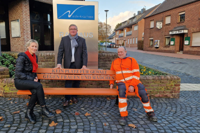 Christiane Jung, Gleichstellungsbeauftragte der Gemeinde Niederkrüchten, Bürgermeister Karl Heinz Wassong und Bernd Pross vom Bauhof der Gemeinde auf einer orangenen Bank mit der Aufschrift "Kein Platz für Gewalt gegen Frauen und Mädchen" vor dem Rathaus in Elmpt