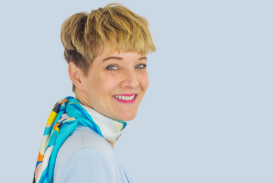 Nadine Diederichs, Expertin auf dem Gebiet der ganzheitlichen Gesundheit, vor einem hellblauen Hintergrund. Sie trägt ein buntes Halstuch und hat den Kopf nach rechts gedreht.