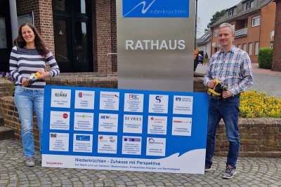 Svenja Sonnemans und Frank Grusen mit einem Veranstaltungsschild vor dem Rathaus in Elmpt. Auf dem Schild die Logos der Unternehmen, die an der Messe "Niederkrüchten - Zuhause mit Perspektive" teilnehmen.
