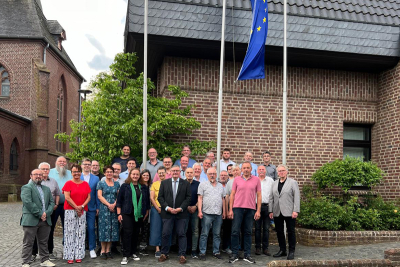Vertreterinnen und Vertreter der Parteien und Wählergemeinschaften im Niederkrüchtener Gemeinderat stehen gemeinsam mit Bürgermeister Karl-Heinz Wassong vor dem Rathaus der Gemeinde Niederkrüchten in Elmpt. Im Hintergrund die Flagge der Europäischen Union.