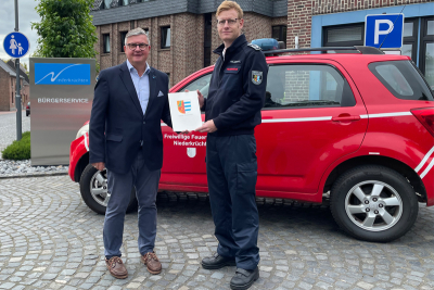 Bürgermeister Karl-Heinz Wassong (links) übergibt die Ernennungsurkunde als stv. Leiter der Feuerwehr an Holger Lütters (rechts). Im Hintergrund ein Feuerwehrfachzeug und ein Schild "Niederkrüchten" vor dem Rathaus der Gemeinde.