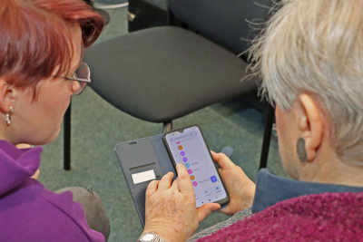 Eine ältere Frau bedient ein Smartphone. Die Person links neben ihr erklärt ihr die Bedienung.