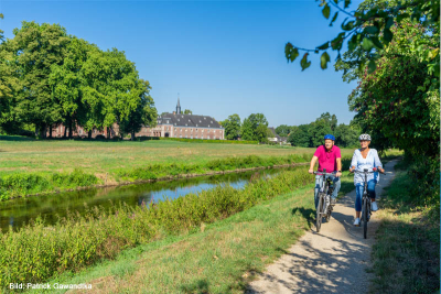 Eine Frau und ein Mann fahren mit dem Fahrrad entlang eines kleinen Flusses. Im Hintergrund ein Schloss.