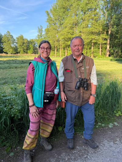 Eine Frau und ein Mann stehen vor einer grünen Wiese und schauen in die Kamera. Im Hintergrund sind grüne Bäume zu sehen.