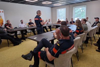 Ein Mann in Kleidung der niederländischen Brandweer redet im Stehen zu mehreren Zuhörenden, die an Tischen sitzen
