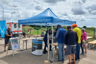 Der Infopunkt der Gemeinde Niederkrüchten beim Raderlebnistag. Ein blauer Pavillon mit Counter, vor dem Teilnehmende der Veranstaltung stehen und sich informieren.