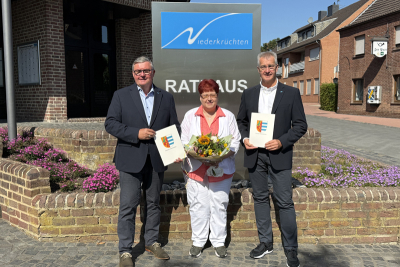 Bürgermeister Karl-Heinz Wassong hält eine Urkunde mit dem Wappen der Gemeinde Niederkrüchten. Neben ihm Silvia Martin und Frank Kriegers. Im Hintergrund ein Schild mit dem Logo der Gemeinde.