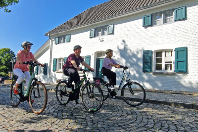 Drei Radfahrende fahren bei gutem Wetter vorbei an "Haus Hansen" in Elmpt.