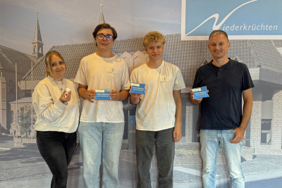 Lea Korall (Klimaschutzmanagerin Gemeinde Niederkrüchten), Merlin Dolenec und Anton Palmer (MerClean) und Frank Grusen (Wirtschaftsförderer Gemeinde Niederkrüchten) vor einer Fotowand mit dem Bild des Rathauses in Elmpt. In den Händen halten Sie Packungen der WC-Tabs
